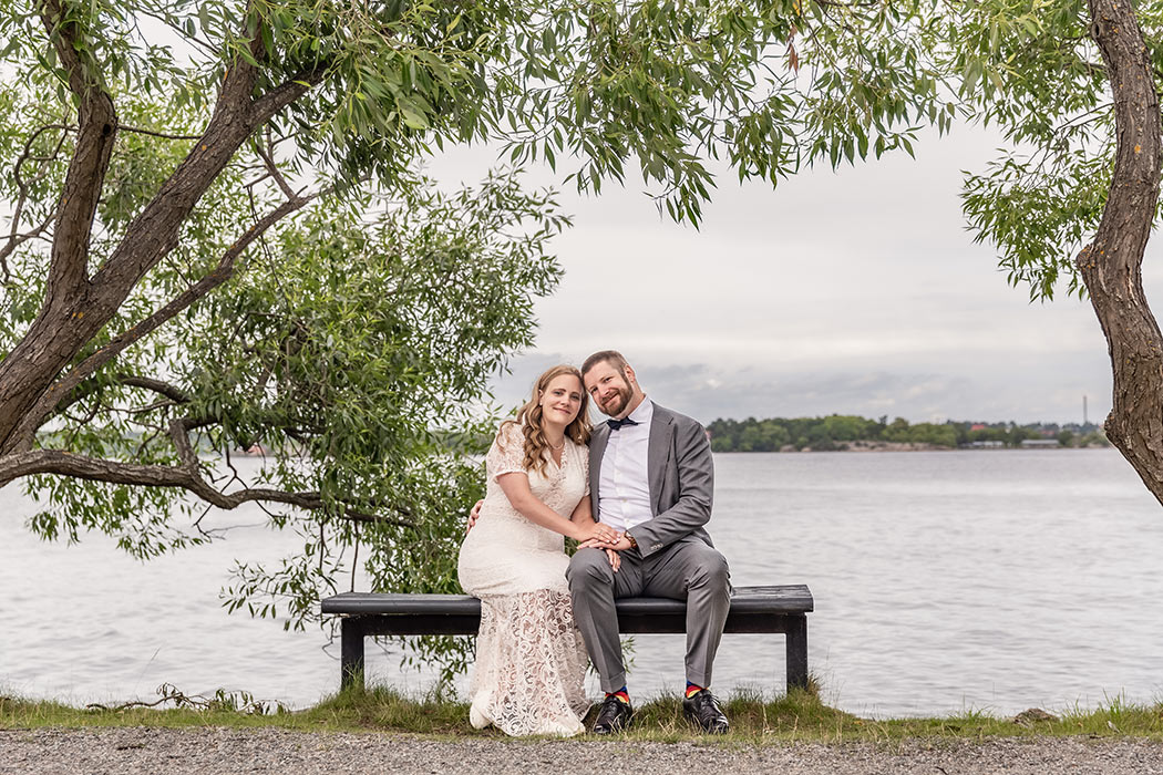 Brudpar sitter på en bänk vid vattenkanten under ett grönskande träd.