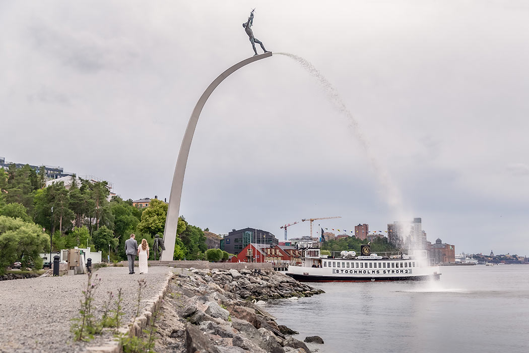 Brudpar går på en grusgång vid vattnet, på väg mot fontänen Gud Fader på Himmelsbågen. I bakgrunden syns Stockholms siluett och en båt.