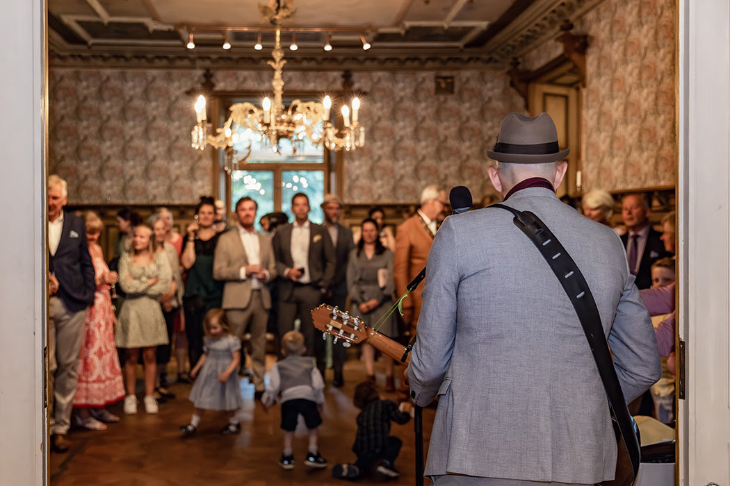 En trubadur spelar på bröllopsminglet i ett gammalt vackert rum. Några små barn springer runt och dansar och leker framför resten av gästerna.