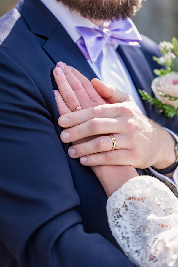Närbild på brudparets händer med ringarna. I bild syns även brudgummens lila fluga.